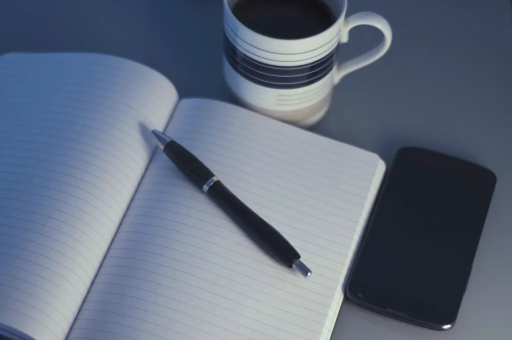 An open notebook, pen, phone and coffee on a table