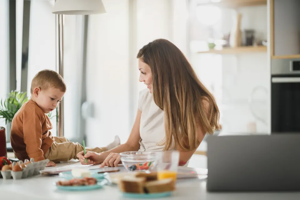 A woman working on a laptop at the kitchen bench while her toddler plays